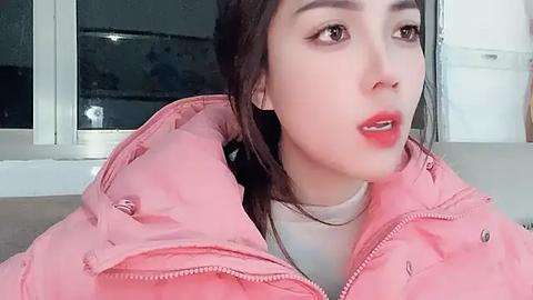 Media: A video of a young Asian woman with fair skin, wearing a pink puffy jacket, red lipstick, and dark hair, looking thoughtful in a modern, minimalist room with white walls and a glass door.