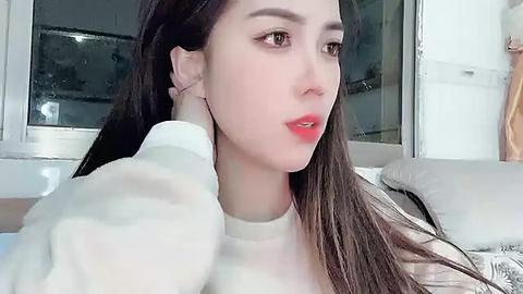 Media: Video of a fair-skinned woman with long, dark hair, wearing a white sweater, sitting indoors near a window. She has a neutral expression, and her left hand is touching her cheek.