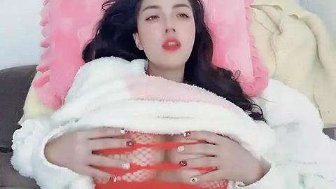 Media: A video of a woman with long dark hair, fair skin, and full red lips, wearing a fluffy white robe and red lingerie, lying on a bed with pink and beige pillows, holding her breasts.