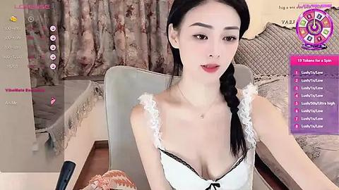 Media: Video of a young Asian woman with light skin, black hair in a braid, wearing a white lace bra, seated on a beige couch in a cozy room.