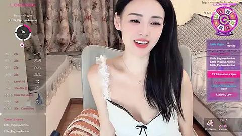 Media: Video of a young, slender Asian woman with long black hair, wearing a white, lace-trimmed camisole, sitting on a bed with floral bedding.