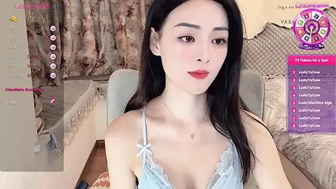 Media: Video of a young East Asian woman with long black hair, fair skin, and red lipstick, wearing a light blue lace bra, seated on a beige couch in a cozy, floral-patterned room.