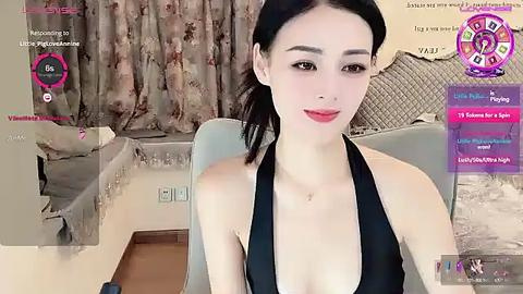 Media: A video of an attractive Asian woman with fair skin, black hair in a ponytail, wearing a black halter top, sitting in a modern living room. The background features beige curtains and a plush sofa.