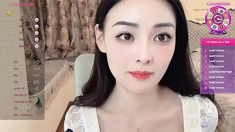 Media: A video of an Asian woman with long, straight black hair, fair skin, and brown eyes, wearing a white lace top. She sits in a room with beige curtains and a purple, circular logo in the corner.