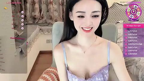 Media: Video of an Asian woman with fair skin, black hair, and a slender build, wearing a lavender lace bra, smiling in a cozy, dimly lit room with floral curtains.