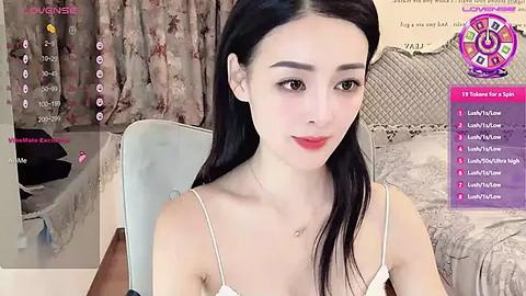 Media: A video of a young East Asian woman with fair skin, long black hair, and a slender physique, wearing a white spaghetti strap top. She sits in a cozy, pink and white room with a floral curtain in the background.