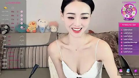 Media: Video of a smiling East Asian woman with light skin and dark hair, wearing a white camisole, seated in a cozy, plush room with stuffed toys.