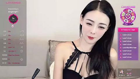 Media: Video of an East Asian woman with long black hair, fair skin, wearing a black lace bra, sitting on a beige couch, with a \"Lovevibe\" screen displaying stats and symbols in the background.