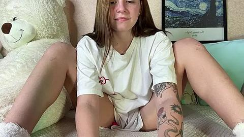 Media: Video of a young woman with light skin, brown hair, and tattoos, wearing a white t-shirt and shorts, sitting on a beige couch with a stuffed bear and a framed Van Gogh painting in the background.
