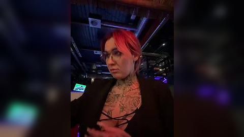 Media: Video of a tattooed woman with red hair, wearing a black top, sitting in a dimly-lit bar with visible neon lights and a wooden ceiling.