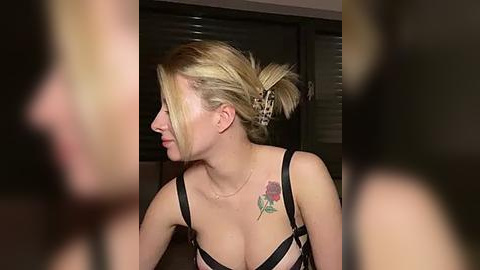 Media: Video of a fair-skinned woman with blonde hair in a ponytail, wearing black lingerie with a floral tattoo on her right shoulder, indoors with dim lighting.