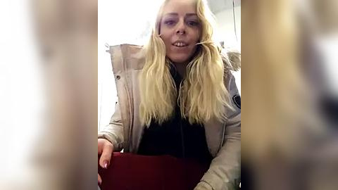 Media: Video of a blonde woman with long, straight hair, wearing a beige jacket and a black top, seated in a train.