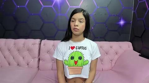 Media: Video of a young Latina woman with straight black hair, medium skin tone, wearing a white T-shirt with a cactus design. She sits on a pink tufted couch against a hexagonal, purple-patterned wall.