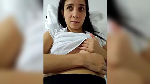 Media: Video of a young woman with medium brown skin and long black hair, lifting a white shirt to expose her small, perky breasts. She lies on a white pillow in a hospital bed, looking concerned.