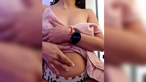 Media: Video of a woman with medium skin tone, wearing a pink off-shoulder top, revealing her bare breasts, and a white skirt with fruit patterns. She has a black smartwatch on her wrist and is holding her pregnant belly.