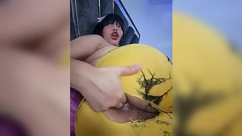 Media: A video of a woman with dark hair, wearing a yellow, torn latex suit, exposing her breasts and genitals. The background shows a blurred, purple room.