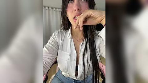 Media: A video of a young woman with long black hair, wearing a white button-down shirt and blue jeans, sitting with her finger in her mouth, blurry background.