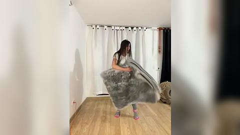 Media: Video of a woman with long brown hair in a gray sweater, holding a fluffy gray blanket, standing on a wooden floor in a minimalist room with white walls and curtains.