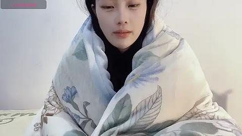 Media: Video of a young Asian woman with long black hair, wearing a white floral-patterned blanket, sitting on a bed with a green and white patterned bedspread, in a softly lit, neutral-toned room.