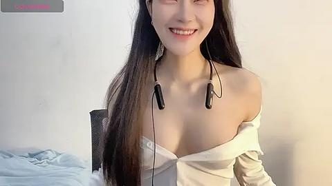 Media: Video of a smiling Asian woman with long black hair, wearing a white off-shoulder top, black headphones, and seated in a chair against a plain white wall.