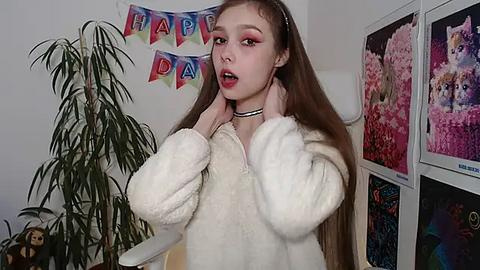 Media: Video of a young woman with long brown hair, wearing a fluffy white robe, standing in a room with \"Happy Birthday\" bunting, a potted plant, and colorful posters on the wall.