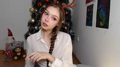 Media: Video of a young Caucasian woman with light skin, brown hair in a braid, wearing a white blouse, red reindeer headband, and sitting indoors next to a decorated Christmas tree with ornaments and a snowman.