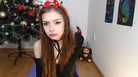 Media: Video of a young woman with long brown hair and red headband, wearing a black choker and black gloves, kneeling on a yoga mat in front of a decorated Christmas tree and wall art in a cozy room.