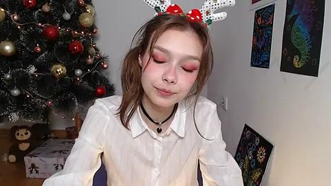 Media: Video of a young woman with fair skin, wearing festive white and red reindeer headband, closed eyes, smiling, in front of a decorated Christmas tree with gold and red ornaments, surrounded by colorful framed art.