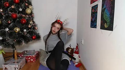 Media: A video of a young woman in a gray dress, black tights, and red antlers, sitting on a purple mat in a Christmas-themed room with a decorated tree, gifts, and colorful artwork.