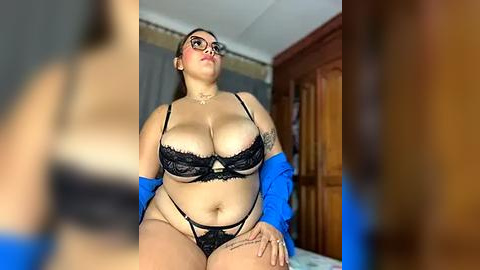 Media: Video of a plus-size Latina woman with a tattoo on her arm, wearing black lace lingerie and a blue robe, sitting on a bed in a dimly lit bedroom.