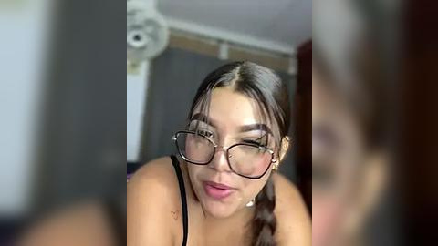 Media: Video of a young woman with light skin, dark hair, and large black-framed glasses, making a playful face with her tongue out. She wears a black tank top.