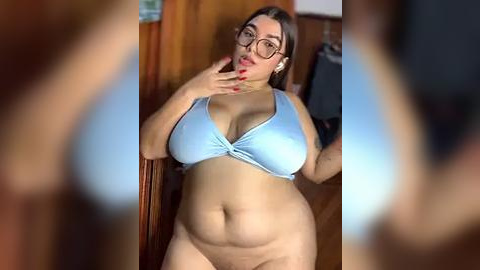 Media: Video of a plus-sized woman with large breasts and wide hips wearing a light blue bikini top and no bottoms. She has medium skin tone, brown hair, and wears glasses. Background includes wooden walls and a blurred figure.