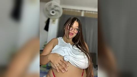 Media: A video of a woman with long brown hair, wearing glasses and a white crop top, lifting it to reveal a red thong, in a dimly-lit room with a ceiling fan.