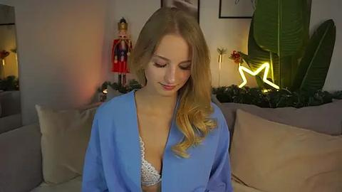 Media: A video of a young, fair-skinned, blonde woman with long hair in a blue robe, sitting on a beige couch, surrounded by Christmas decorations and a glowing neon star.