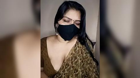 Media: Video of a South Asian woman with long black hair, wearing a black face mask, a beige saree with a deep neckline, and dangling earrings.