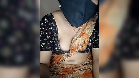 Media: Video of a woman wearing a floral-patterned saree, black blouse, and blue mask, with a blurred background.