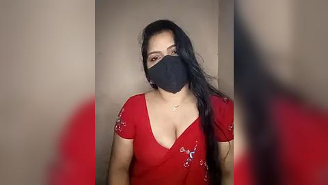 Media: Video of a young woman with long black hair, wearing a red dress, black face mask, and a gold necklace, against a beige background.