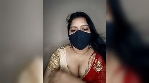 Media: Video of a woman with long black hair, wearing a red and gold sari, black face mask, and large earrings, against a blurred background.