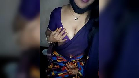 Media: A dimly-lit video of a woman with medium skin tone, wearing a purple blouse and colorful skirt, lifting her blouse to reveal her cleavage. She has a black choker necklace and long black hair.