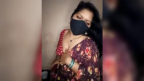 Media: Video of a South Asian woman with long black hair, wearing a floral-patterned sari and black face mask, with a blurred background.