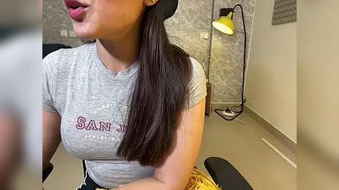 Media: Video of a Latina woman with long, dark hair, wearing a tight grey \"Santana\" t-shirt, sitting in a salon chair, with a yellow lamp and grey patterned wall in the background.