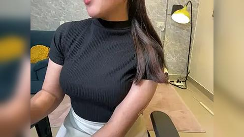 Media: Video of a woman with straight, dark hair, wearing a black ribbed crop top and light blue skirt, seated in a modern living room with a yellow lamp and grey walls.