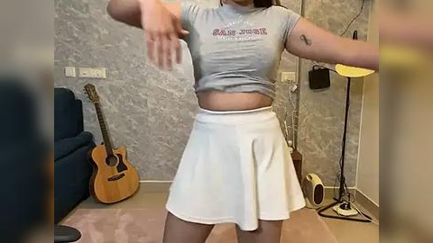 Media: Video of a woman in a gray crop top and white skirt, mid-dance, in a room with a blue couch, guitars, and a lamp.