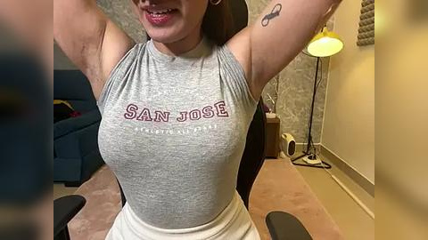 Media: Video of a smiling woman with light skin, wearing a tight, gray San Jose \"The City\" tank top, revealing her medium-sized breasts, and a white skirt, in a modern, beige-walled room with a lamp and a chair.