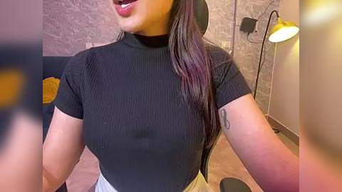 Media: Video of a light-skinned woman with long, dark hair, wearing a black ribbed short-sleeve top and white pants, smiling, in a modern living room with a textured grey wall, a yellow lamp, and a dark blue couch.