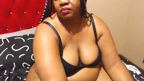 Media: Video of a plus-size Black woman with braids, wearing a black bra, sitting on a bed with a red tufted headboard and black and white patterned sheets.