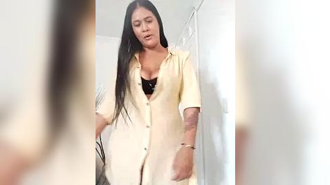 Media: Video of a woman with long black hair, wearing a loose, yellow button-down shirt, partially revealing a black bra. She stands in a brightly lit room with white walls and a shower curtain in the background.