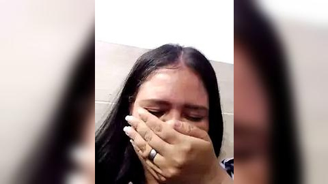 Media: Video of a woman with long black hair, covering her face with her hands, wearing a silver ring, in a bathroom with beige tiles.