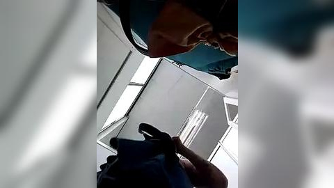 Media: A video capturing a person's head and torso inside a small, dimly lit room with a blue backpack, wearing a light brown shirt. The background shows a white ceiling with fluorescent lighting.