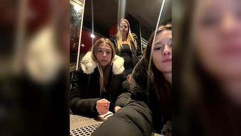 Media: Video of three young women in a dimly lit subway car, wearing winter coats with fur linings. One woman holds a phone, another leans forward, and the third stands behind them.